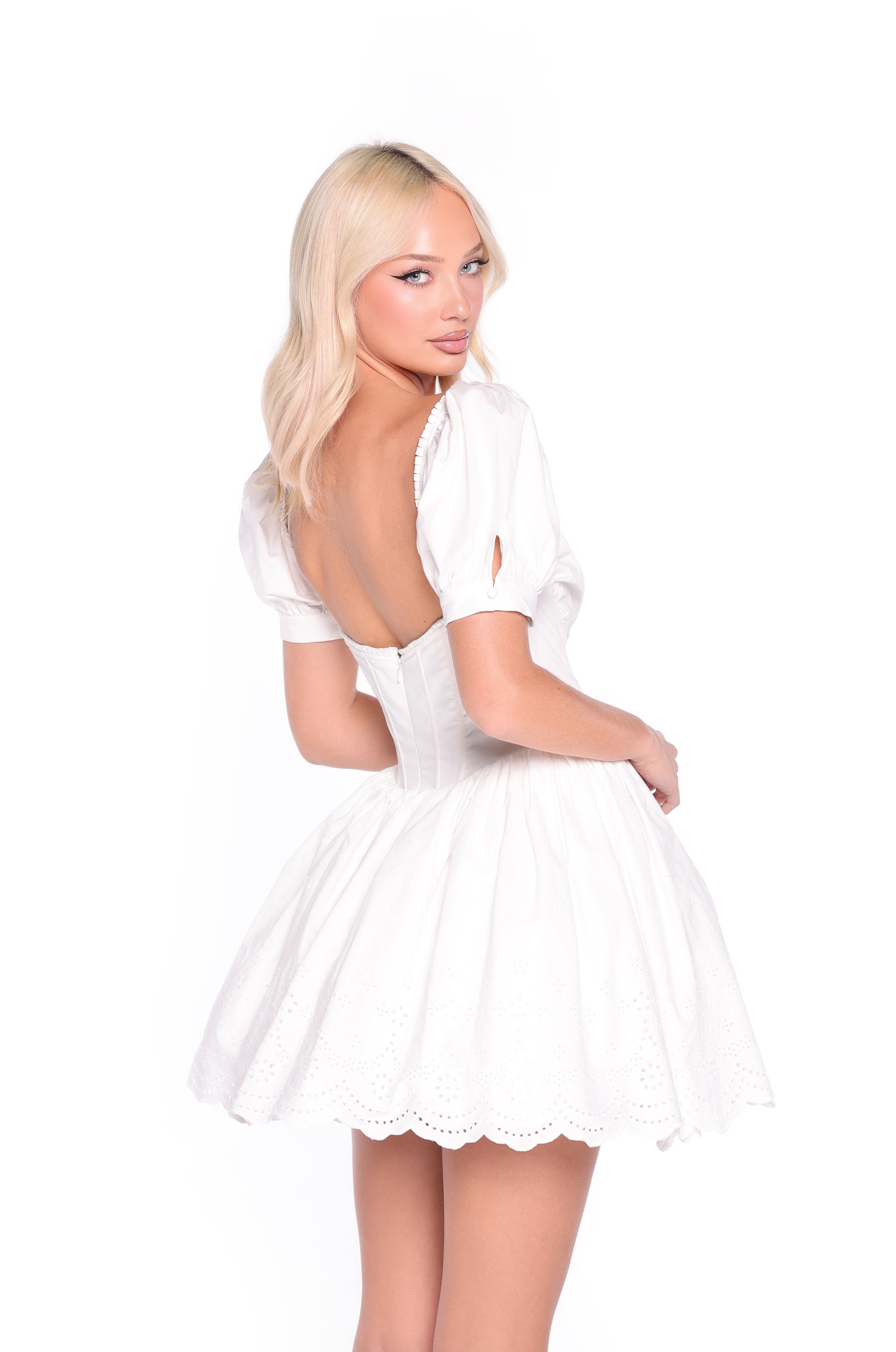 A woman with long blonde hair models the Celine Fit-and-Flare Short White Grad Dress, featuring a low back and puffed sleeves, against a plain white background.